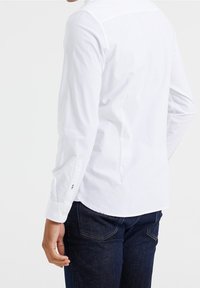 Biała koszula z długim rękawem, wyposażona w kołnierzyk zapinany na guziki, mankiety na pojedynczy guzik oraz zaokrąglony dół, noszona z ciemnoniebieskimi dżinsami. Gładka, bawełniana faktura.