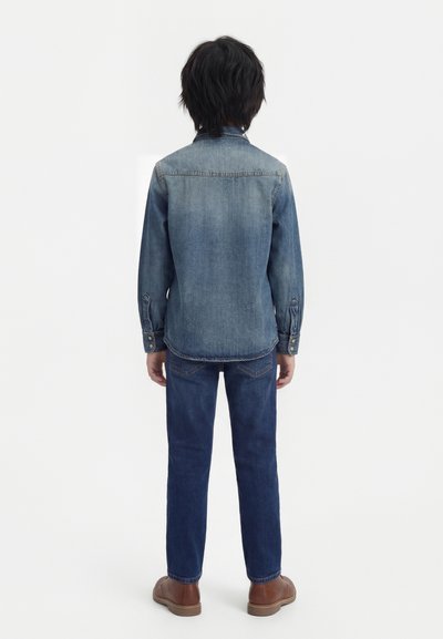 Παιδί με μαύρα μαλλιά που φοράει ξεθωριασμένο τζιν μπουφάν, σκούρο μπλε τζιν και καφέ παπούτσια, στέκεται γυρισμένο με την πλάτη σε απλό φόντο.