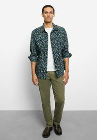 Marineblaues Hemd mit Blumenmuster und hochgekrempelten Ärmeln, kombiniert mit olivgrünen Hosen. Modell steht auf grauem Boden, trägt dunkle Schuhe und weißes Hemd.