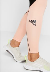 Světlé růžové sportovní legíny z elastického materiálu s černým logem Adidas a kontrastními černými bočními švy. Kombinovány s světle šedými teniskami.