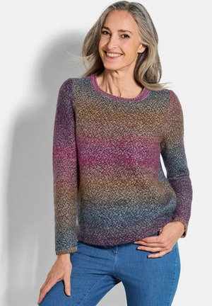 Eine mittelalte Frau mit grauem Haar lächelt, trägt einen mehrfarbigen Strickpullover und eine bluejeans und steht vor einem schlichten hellen Hintergrund.