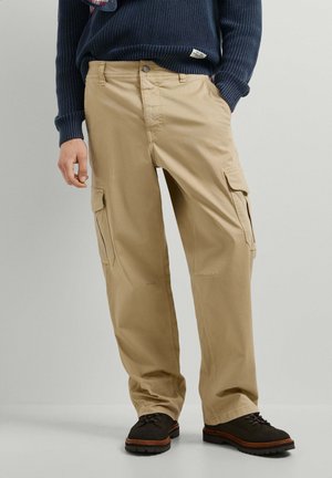 Mann trägt beige Cargo-Hose, schwarze Schuhe und einen dunkelblauen Pullover mit den Händen in den Taschen vor einem einfarbigen Hintergrund.