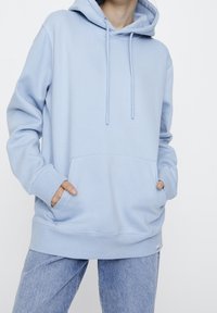 Personne portant un sweat à capuche bleu clair avec les mains dans la poche avant, associé à un jean denim bleu clair, devant un fond blanc uni.