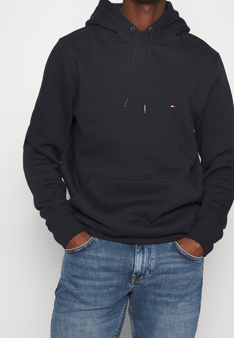 Sudadera azul marino hecha de mezcla de algodón, con un bolsillo tipo canguro, capucha con cordón y pequeño logo en el pecho. Combinada con vaqueros azules.