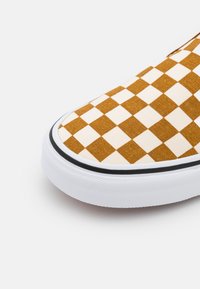 Bruna och krämfärgade rutiga kanvas slip-on skor med en vit gummisula och svart kant, med en texturerad tygyta.