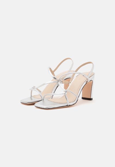 Paire de sandales argentées à brides avec talons hauts carrés et talons blocs sur un fond clair uni.