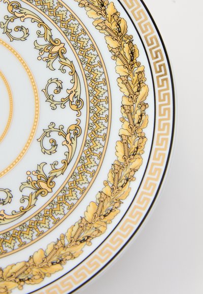 Porcelāna šķīvis ar zelta un melniem sarežģītiem ziedu un grieķu atslēgas rakstiem. Apaļas formas, ar gludu, spīdīgu balto virsmu.