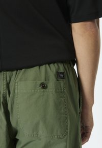 Pantalones cortos cargo verdes con cintura ajustable de cordón, que incluyen un bolsillo trasero con botón y un pequeño parche con logo. La tela parece suave y ligera.
