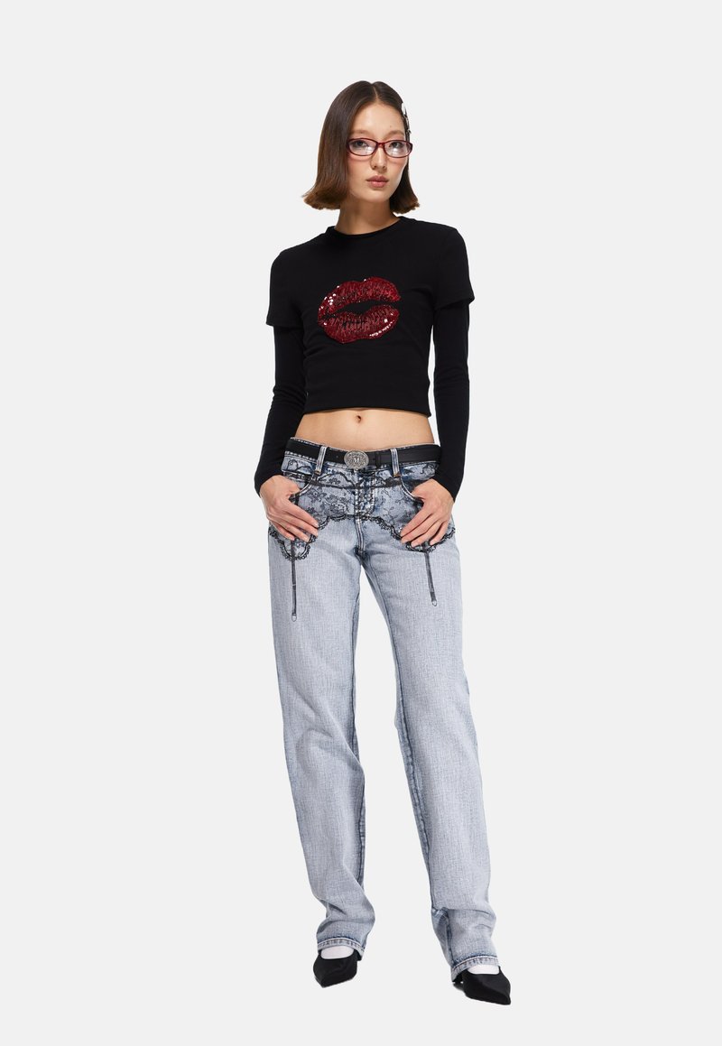 Top corto negro con gráfico de labios de lentejuelas rojas, combinado con jeans de pierna ancha azul claro que presentan costuras decorativas y cremalleras. Incluye un cinturón negro.