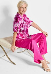 Rosa Blumenoberteil mit fließenden Ärmeln, kombiniert mit einfarbigen rosa Hosen. Das Model sitzt auf einer hellen, plüschigen Bank und trägt beigere hohe Sandalen.