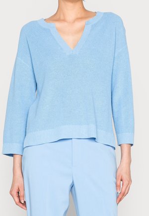 Lyseblå V-hals strikket trøje med ribstrikkede manchetter og kant, kombineret med lyseblå bukser på en stående person beskåret ved halsen.