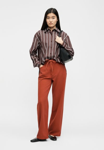 Νεαρή γυναίκα με μακριά μαύρα μαλλιά, φορώντας ένα καφέ και λευκό ριγέ πουκάμισο, παντελόνι σε χρώμα σκουριάς, μαύρα παπούτσια και κρατώντας μια μαύρη τσάντα χειρός.