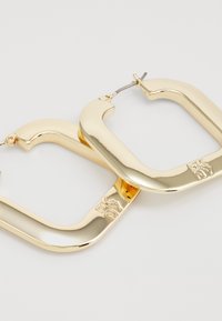 Orecchini a cerchio oro con design a forma di U, finitura lucida e dettaglio del marchio DKNY sulla parte inferiore.