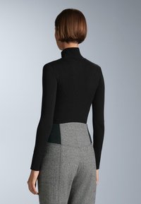 Čierny rebrovaný rolák v kombinácii so šedými štruktúrovanými nohavicami s vysokým pásom, ktoré majú tmavozelené akcenty na páse.