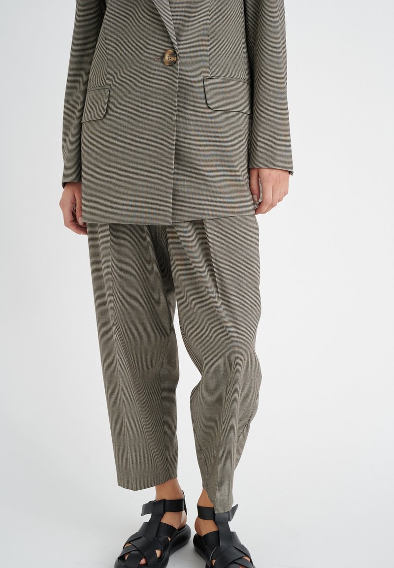 Blazer gris à rayures avec fermeture à bouton unique, assorti à un pantalon gris ample. Le matériau semble texturé. Des sandales noires à lanières complètent le look.