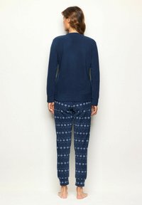 Mujer de pie descalza, de frente a la pared, vestida con una blusa de manga larga azul marino y pantalones de pijama azul marino con estampado.
