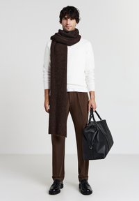 Ein brauner Strickschal liegt über einem weißen Pullover; kombiniert mit braunen Hosen und schwarzen Schuhen. Eine schwarze Lederhandtasche wird in einer Hand gehalten.