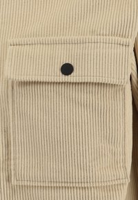 Tessuto in corduroy beige con righe verticali, dotato di una tasca applicata con chiusura a scatto nera. La texture è morbida e strutturata.