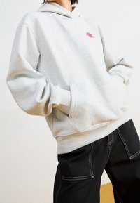 Persona che indossa un hoodie grigio chiaro con logo rosso e pantaloni neri con cuciture bianche, mani nelle tasche dell'hoodie, su uno sfondo leggero e uniforme.