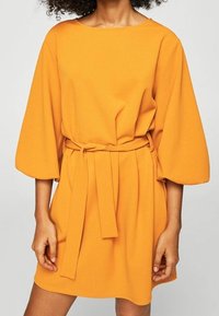 Pomarańczowa sukienka z paskiem, okrągłym dekoltem i rękawami o długości trzech ćwiartek, uszyta z miękkiego materiału o gładkiej fakturze.