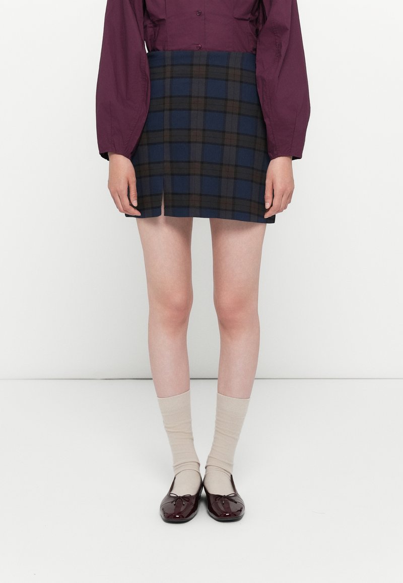Geruite mini-rok met een blauw en bruin patroon, met een rechte snit en een voorknip, gecombineerd met beige sokken en glanzende bordeauxrode schoenen.