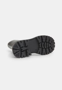 Suela de bota de goma negra con un patrón de banda de rodadura profundo y agresivo, diseñado para ofrecer tracción. Superficie superior suave con forma redondeada.