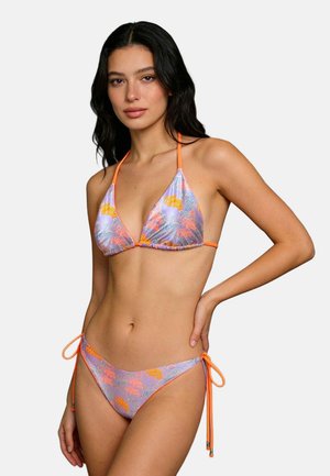 Žena s dlhými tmavými vlasmi má na sebe pastelovo fialové a oranžové kvetinové bikiny s viazaním za krk a nohavičkami na zaväzovanie na bokoch, stojí pred jednoduchým pozadím.