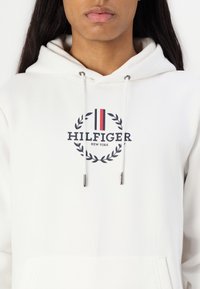 Hvit hettegenser med en marineblå logo med teksten "HILFIGER" og "NEW YORK", rammet inn av en laurbærkrans, og sølvfargede snorer.