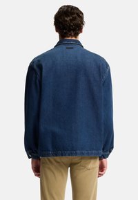 Giacca di jeans blu scuro, con colletto classico, maniche lunghe e vestibilità leggermente rilassata. Nessun logo visibile.