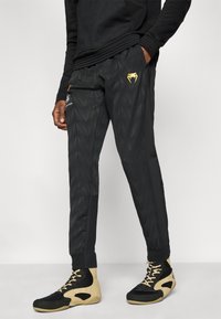 Μαύρο αθλητικό παντελόνι με υφασμένο τρίγωνο μοτίβο, κίτρινες λεπτομέρειες και πλαϊνές τσέπες, σε συνδυασμό με μαύρα και καφέ αθλητικά παπούτσια.