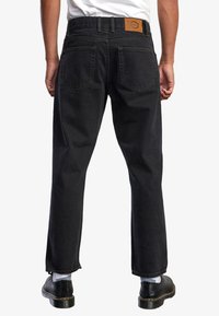 Jeans de mezclilla negra con corte recto, que presentan un parche de cuero marrón en la cinturilla trasera y dos bolsillos traseros.