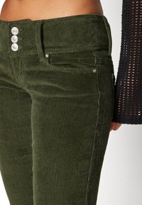 Zelenohnědé kordové kalhoty s třemi stříbrnými knoflíky na vysokém pasu, s vertikálním strukturovaným vzorem a předními kapsami.