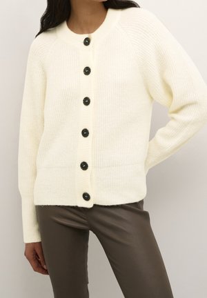Cardigan blanc en tricot avec six boutons foncés, porté avec un pantalon en cuir marron, main du mannequin posée sur la hanche, torse et partie supérieure des jambes visibles.