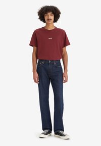 Mlad muž s kovrdžavom kosom koji nosi tamnocrvenu Levi's majicu, tamnoplave traperice i crne tenisice na bijeloj pozadini.