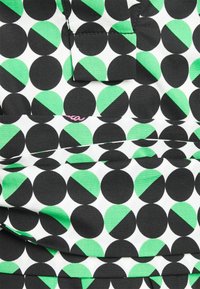 Mönstrad tyg med växlande svarta och gröna cirklar på en vit bakgrund, med en rufsig textur och en liten rosa logotypdetalj.