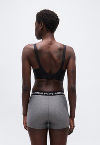 Γυναίκα με κοντά μα λλιά, φοράει μαύρο αθλητικό σουτιέν και γκρι σορτς Under Armour, στέκεται με την πλάτη στην κάμερα μπροστά σε απλό φόντο.