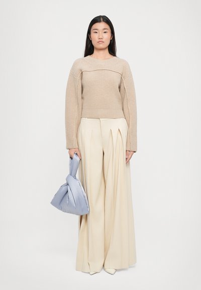Maglione beige a maglia con texture a coste, abbinato a pantaloni a vita alta plissettati color crema. Tiene in mano una borsa annodata di colore azzurro chiaro.