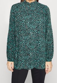 Kragblus med långa ärmar i teal med ett svart leopardmönster, med en knappslå och lätt utsvängd nederkant.