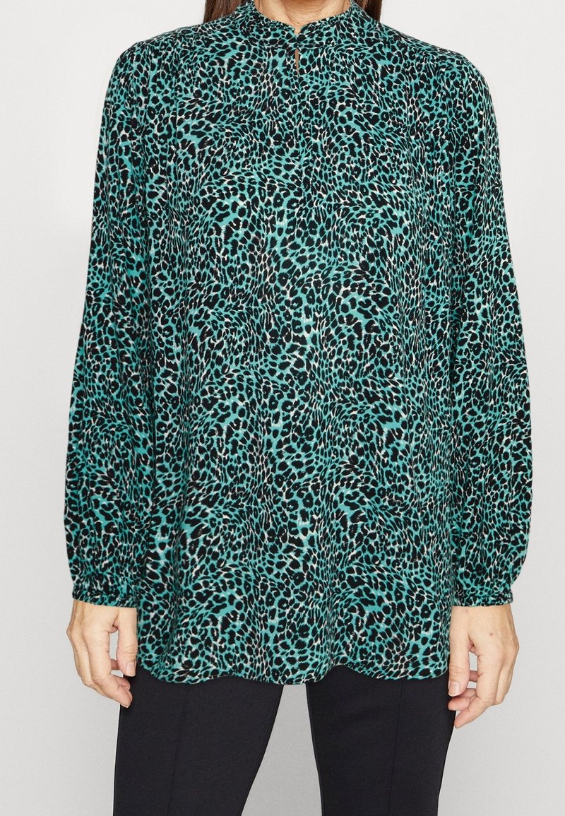 Kragblus med långa ärmar i teal med ett svart leopardmönster, med en knappslå och lätt utsvängd nederkant.