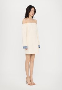 Rochie crem, cu umerii expuși, cu mâneci lungi și accente de denim la manșete. Textură fină, croială până la genunchi, purtată cu sandale bej cu bretele.