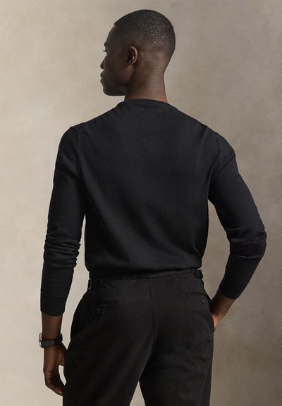 Mann, von hinten gesehen, trägt ein eng anliegendes schwarzes Langarmshirt und schwarze Hose vor einem einfarbigen beigen Hintergrund.