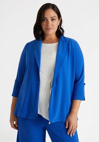 Niebieski żakiet z teksturowanej tkaniny, zapinany na jeden guzik, z rękawami o długości trzech ćwiartek. Nosi się go na białej bluzce z detalem na sznurku.