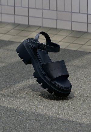 Sandalo nero con suola spessa, ampie fasce e cinturino con fibbia alla caviglia su marciapiede grigio vicino a muro piastrellato.