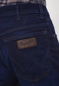 Mörkblå jeans i denim med en läderlapp från Wrangler på bakfickan, med zickzack-sömmar och en slät textur.
