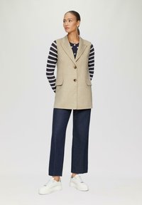 Beige Wolle Weste mit taillierter Passform, ausgestattet mit zwei frontalen Taschen und einem Knopfverschluss, kombiniert mit einem navy gestreiften Langarmoberteil und dunklen Jeans.