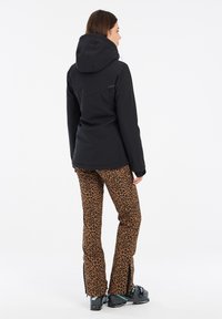 Schwarze wasserdichte Jacke mit Kapuze, kombiniert mit braunen Leopardenmuster-Hosen mit Reißverschluss am Saum und schwarzen Skistiefeln.