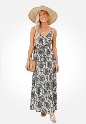 Femme souriante portant un chapeau de paille à large bord, une robe maxi à motifs noirs et beiges, un sac bandoulière tissé et des sandales blanches.