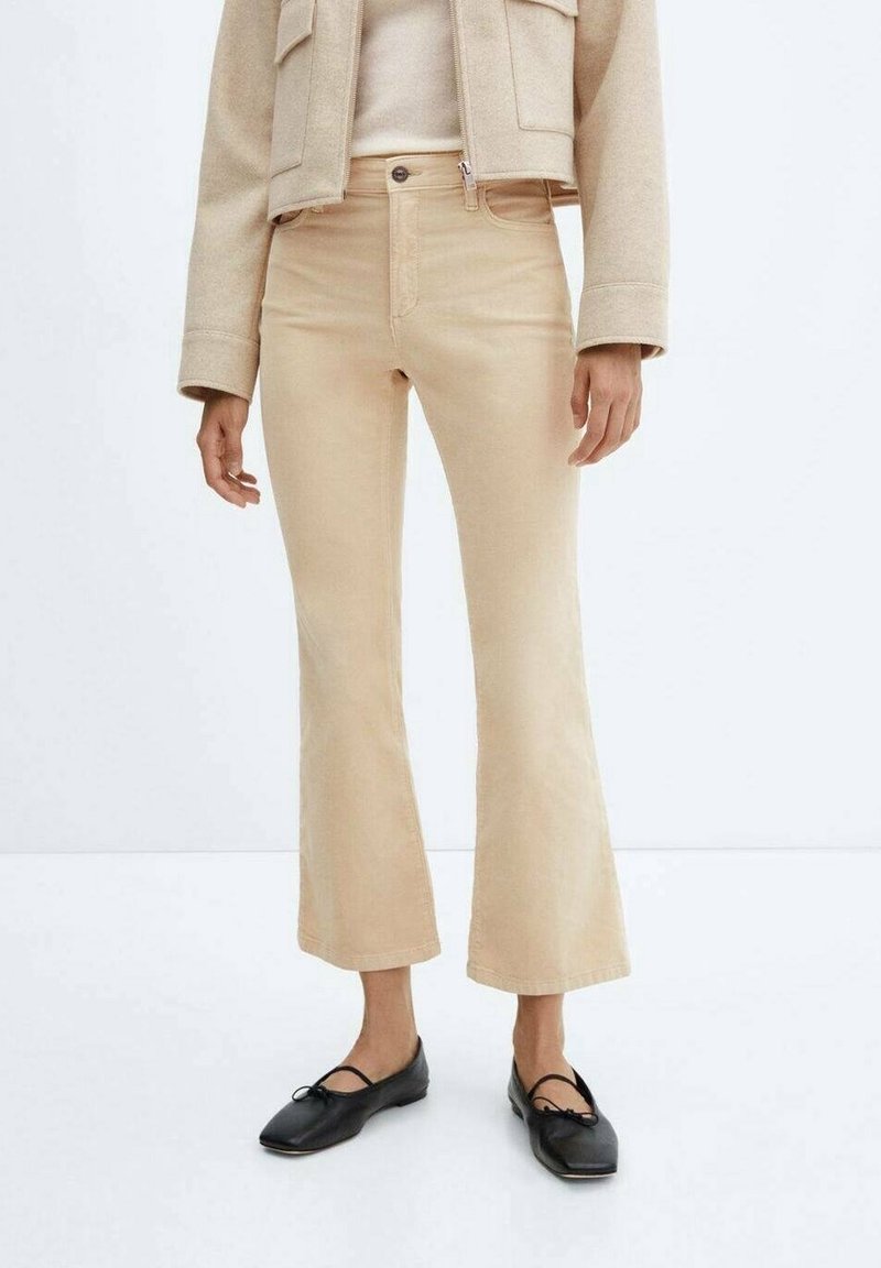Pantalon flare beige raccourci porté avec une veste beige et des ballerines noires à bout carré sur un fond blanc.