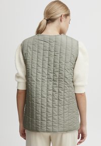 Vadderad väst i dämpad olivgrön, med vertikal sömnad, ärmlös design och rundad fåll. Texturerad krämfärgad tröja synlig under.
