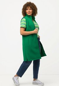 Grüne ärmellose Weste mit hohem Kragen, mit Reißverschluss und Seitentaschen, kombiniert mit einem gestreiften T-Shirt und blauen Jeans, getragen mit weißen Sneakern.
